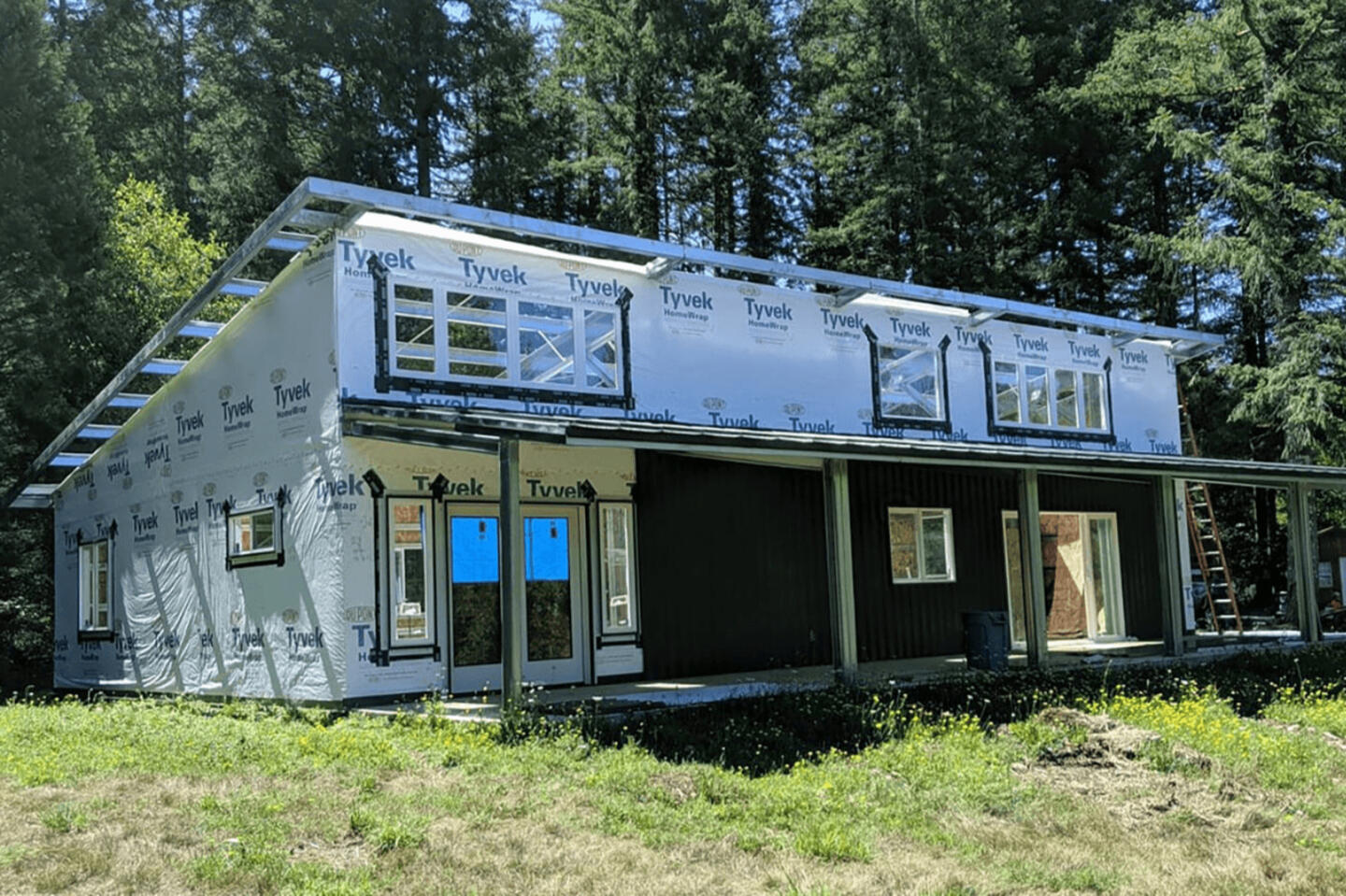 Two-story steel kit home under construction wrapped in Tyvek HomeWrap house wrap with installed windows, surrounded by tall trees